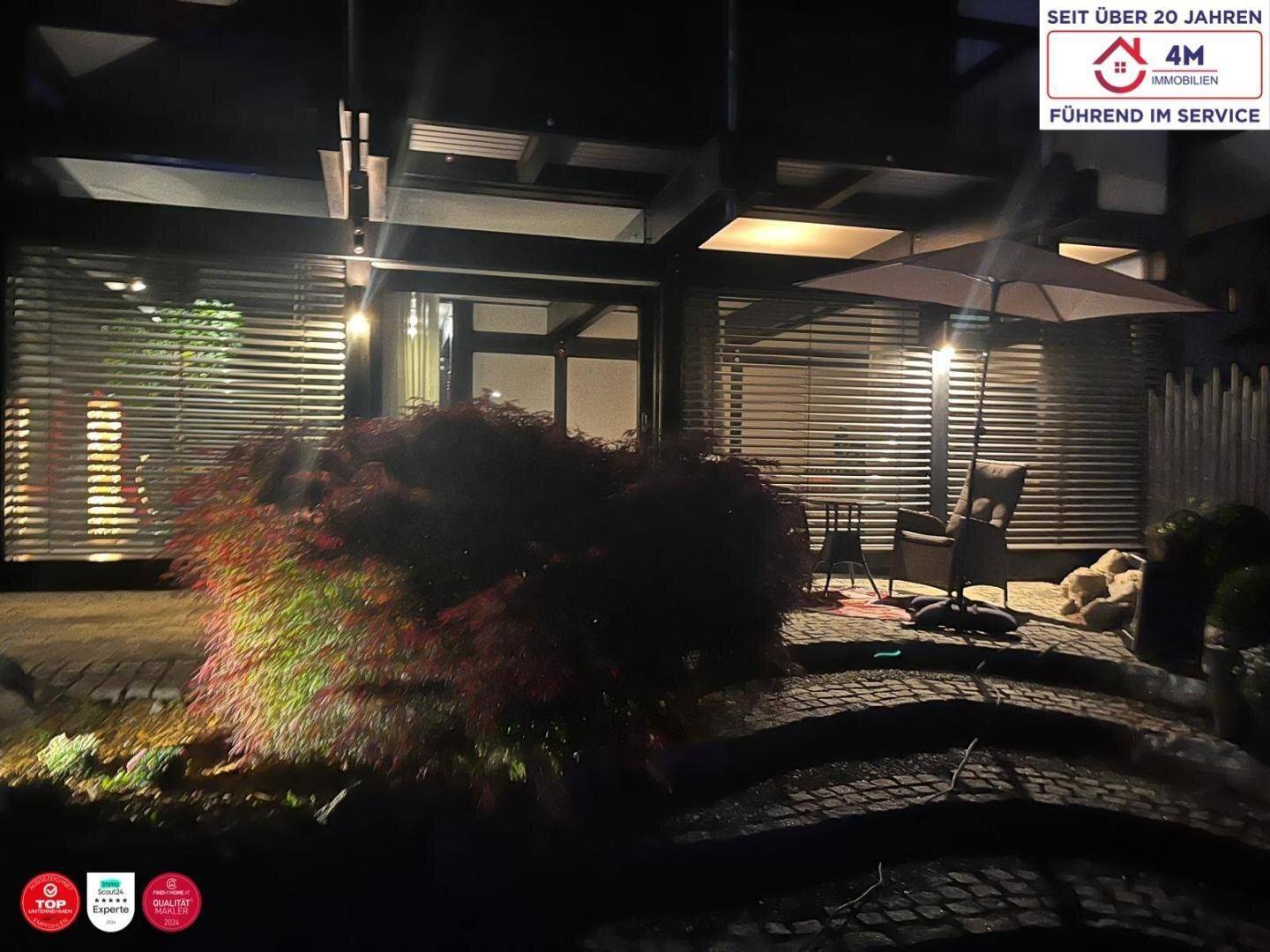 Beleuchtete Terrasse am Abend mit Gartenmöbeln und einem dekorativen roten Strauch im Vordergrund.