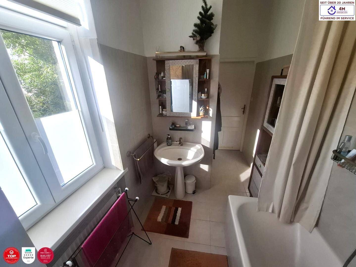 Helles Badezimmer mit Fenster, Badewanne, Waschbecken und Spiegelschrank.