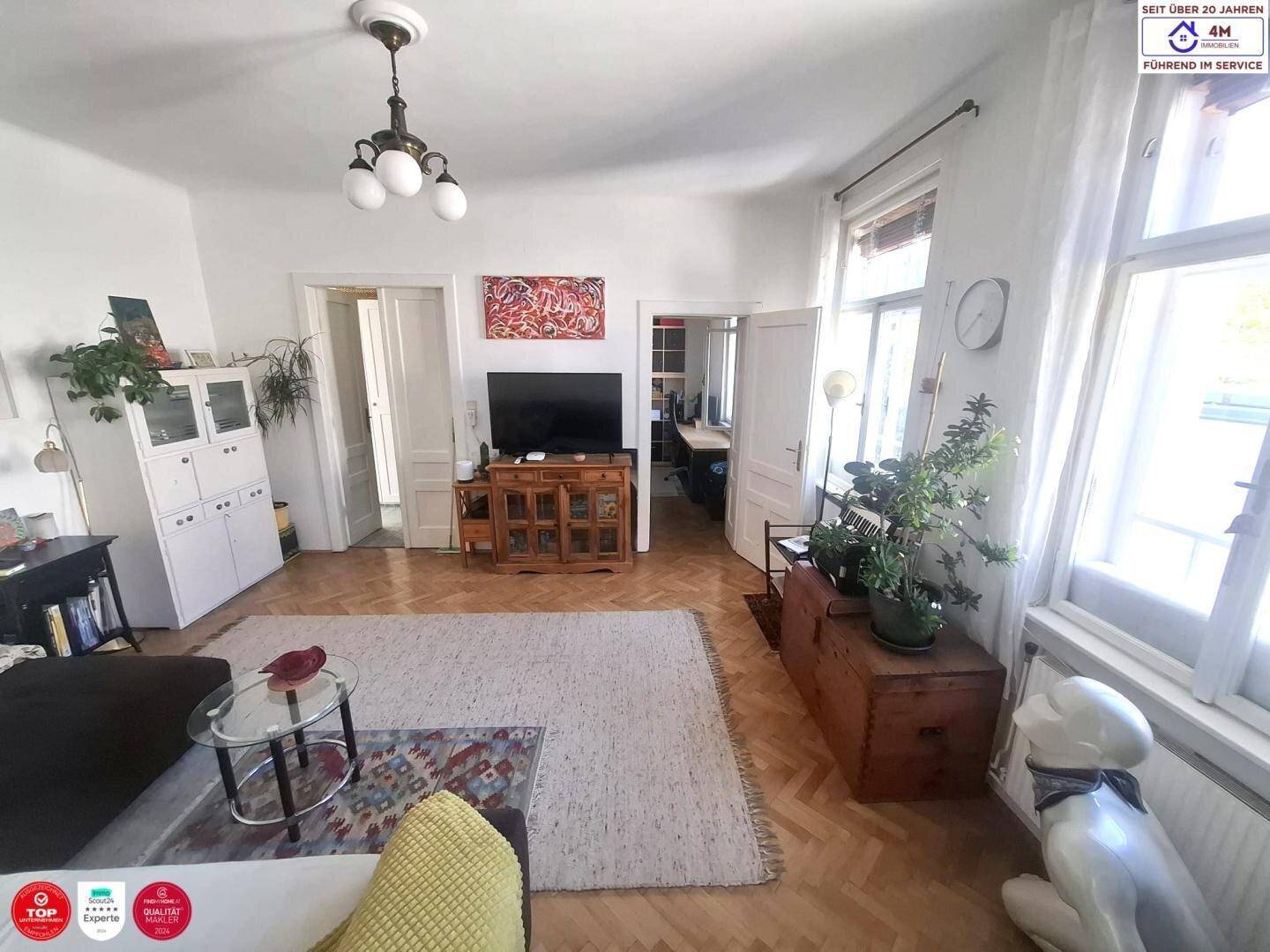 Lichtdurchflutetes Wohnzimmer mit Parkettboden, Fernseher und Zugang zu weiteren Räumen.