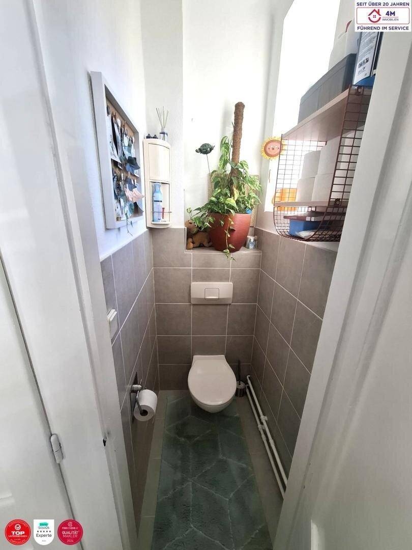 Separates WC mit Wand-WC, Fliesen und kleinem Waschbecken, gut beleuchtet.