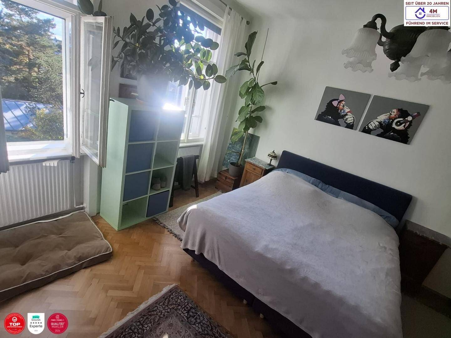 Schlafzimmer mit Doppelbett, Parkettboden und großen Fenstern, die viel Licht hereinlassen.