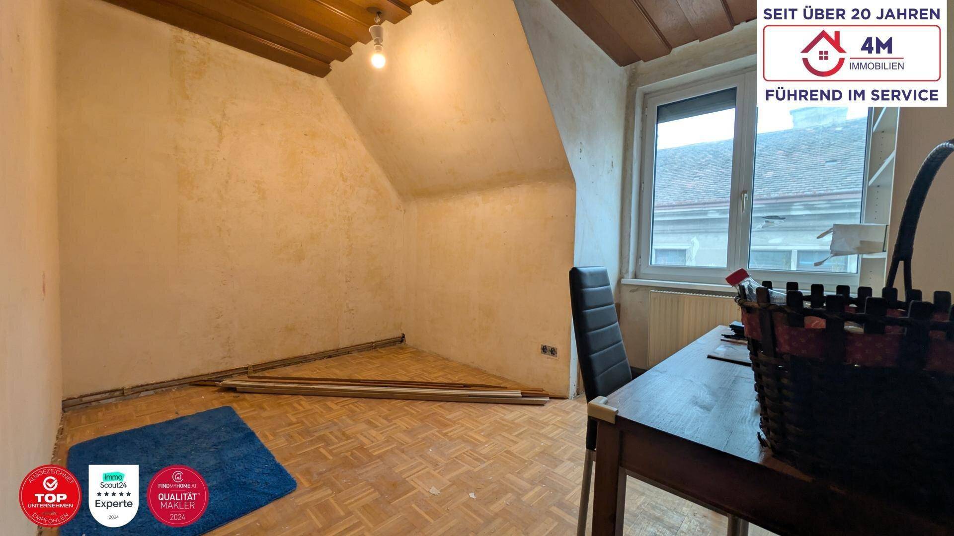 Unrenoviertes Zimmer mit Dachschräge, Parkettboden und Blick aus dem Fenster.