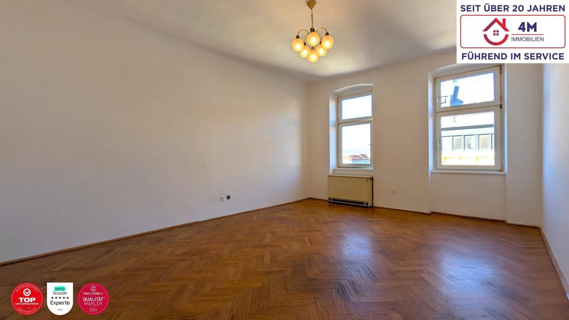 Geräumiger, unmöblierter Raum mit Fischgrätparkett, hohen Decken und großen Fenstern.