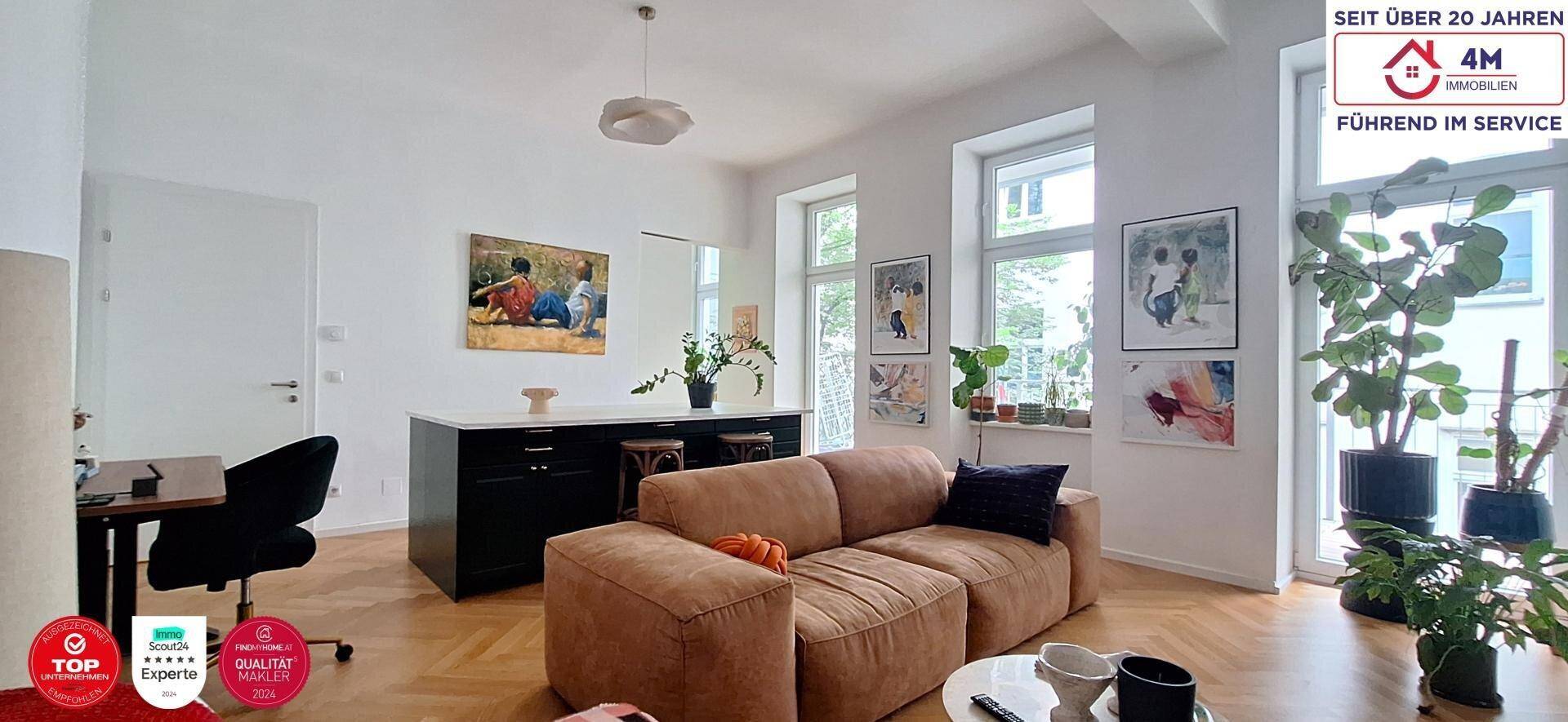 Helles Wohnzimmer mit beigem Sofa, Schreibtisch und vielen Pflanzen, offen zur Küche.