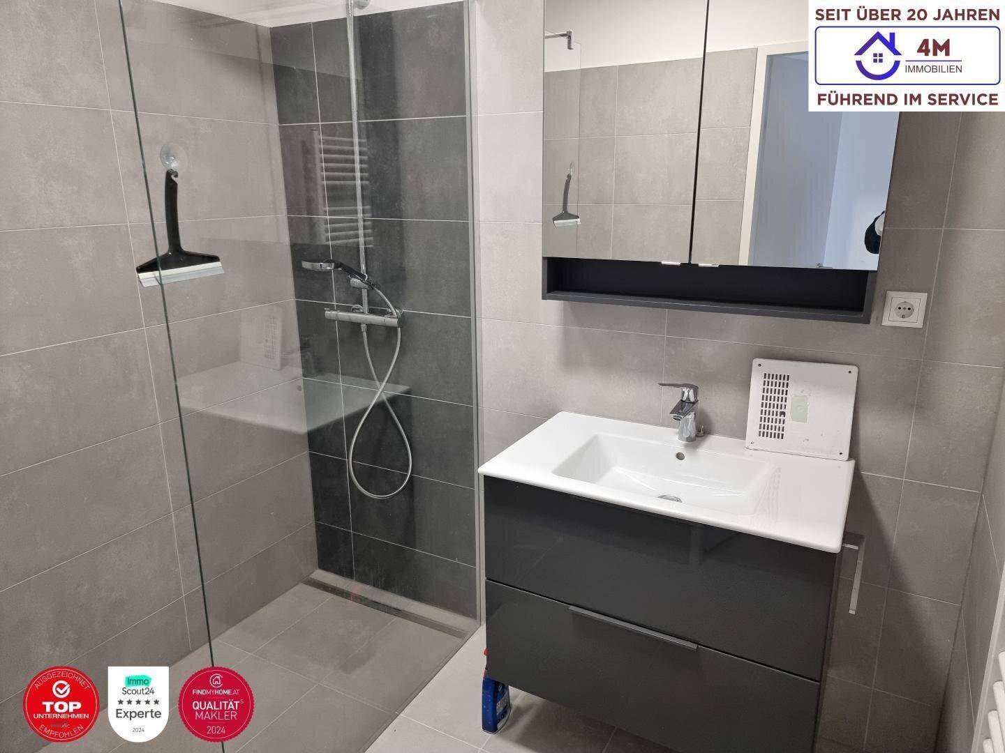 Modernes Badezimmer mit grauen Fliesen, Duschkabine und einem einzelnen Waschbecken mit Unterschrank.