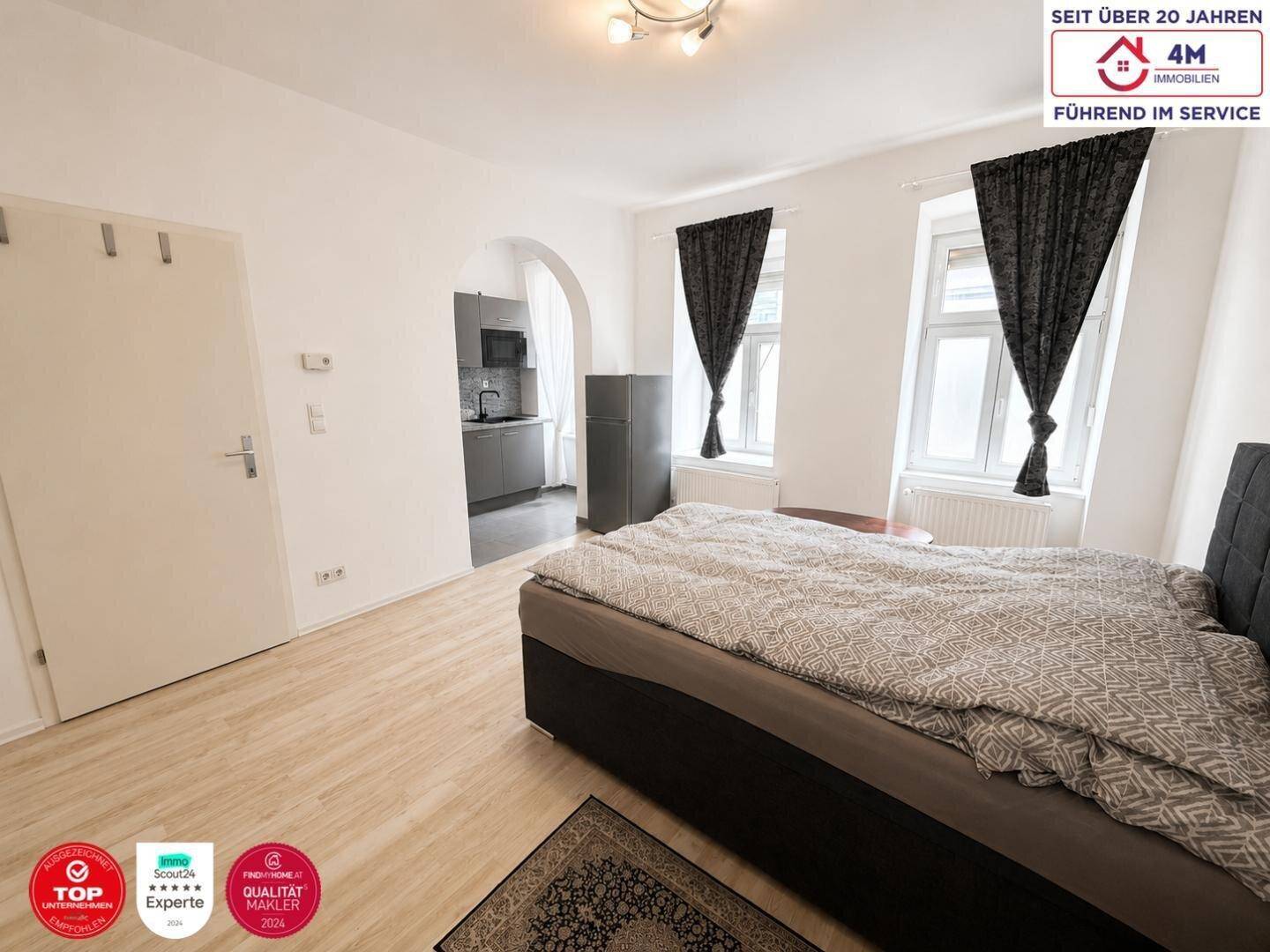 Geräumiges Schlafzimmer mit Bett, Fenstern und offenem Zugang zur Küchenzeile durch einen Rundbogen.