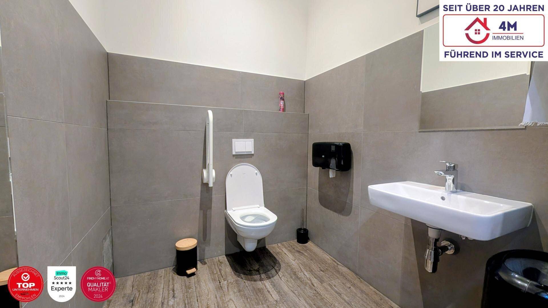 Barrierefreies Badezimmer mit Wand-WC, Haltegriffen und modernem Waschbecken in Grautönen.