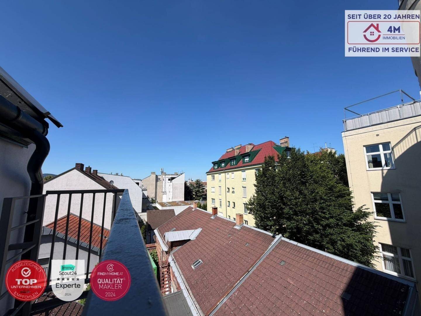 Weitblick vom Balkon über die Dächer der Stadt und grüne Bäume unter blauem Himmel.