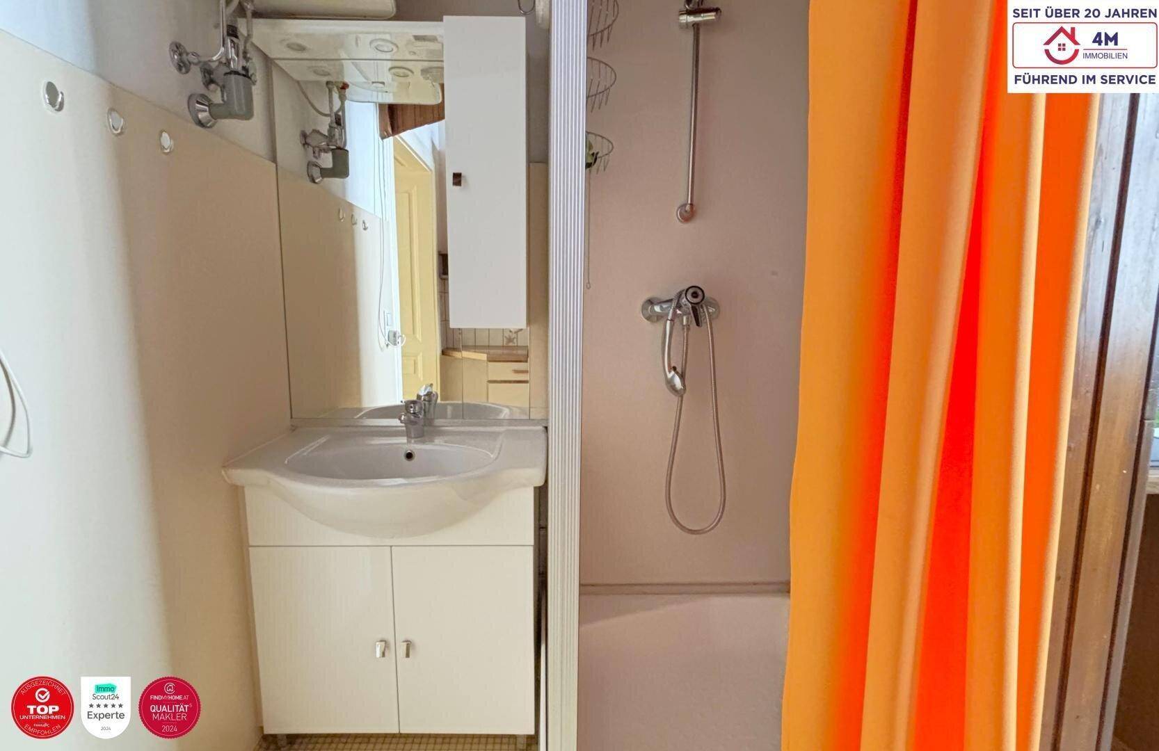 Kleines Badezimmer mit Waschbecken, Spiegel und Duschbereich mit orangefarbenem Vorhang.