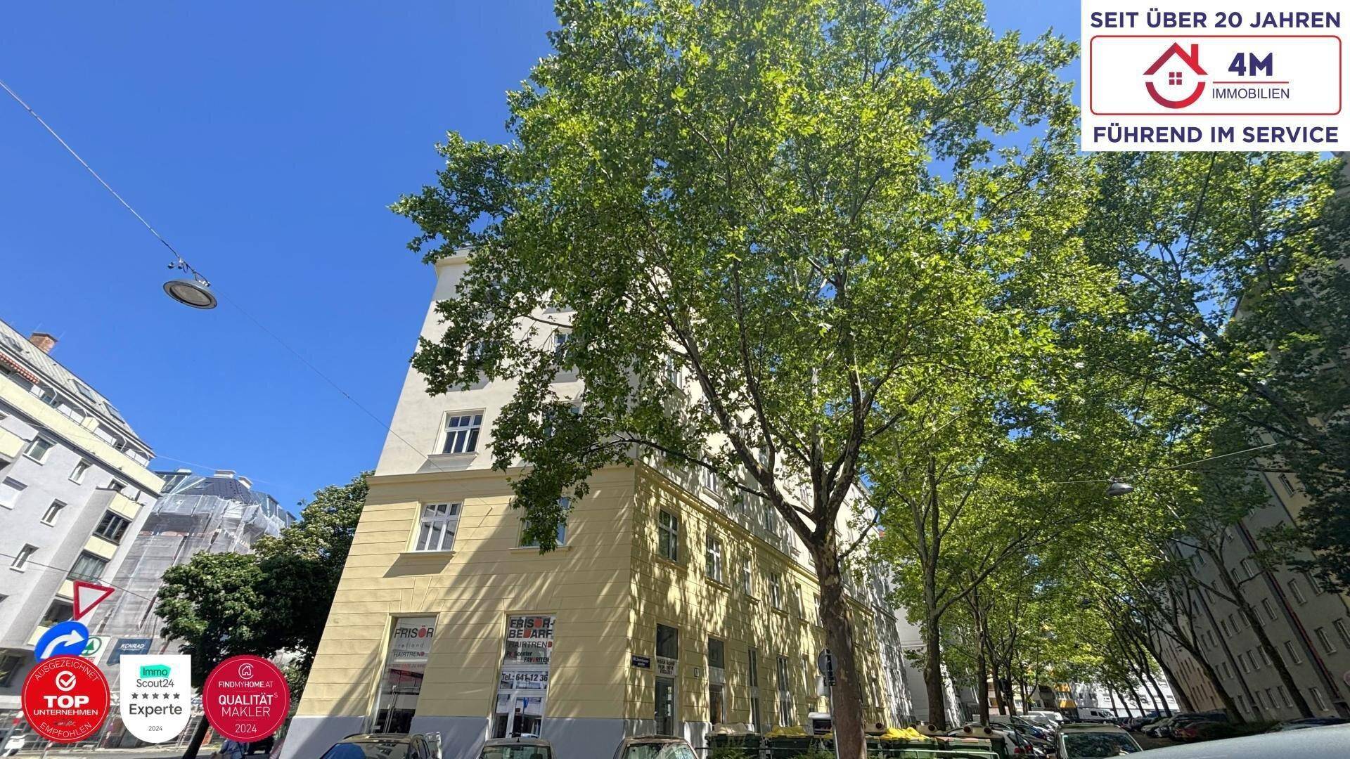 Historisches Gebäude mit hohen Bäumen und blauem Himmel in einer städtischen Umgebung.