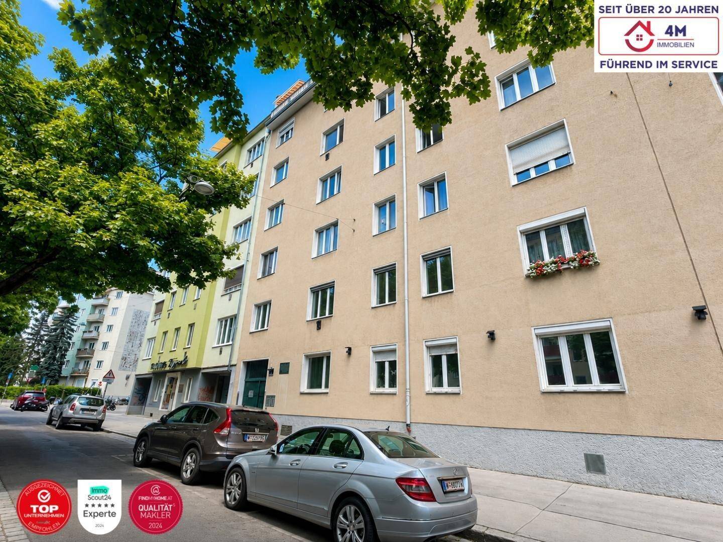 Mehrfamilienhausfassade an einer städtischen Straße mit parkenden Autos und Bäumen.