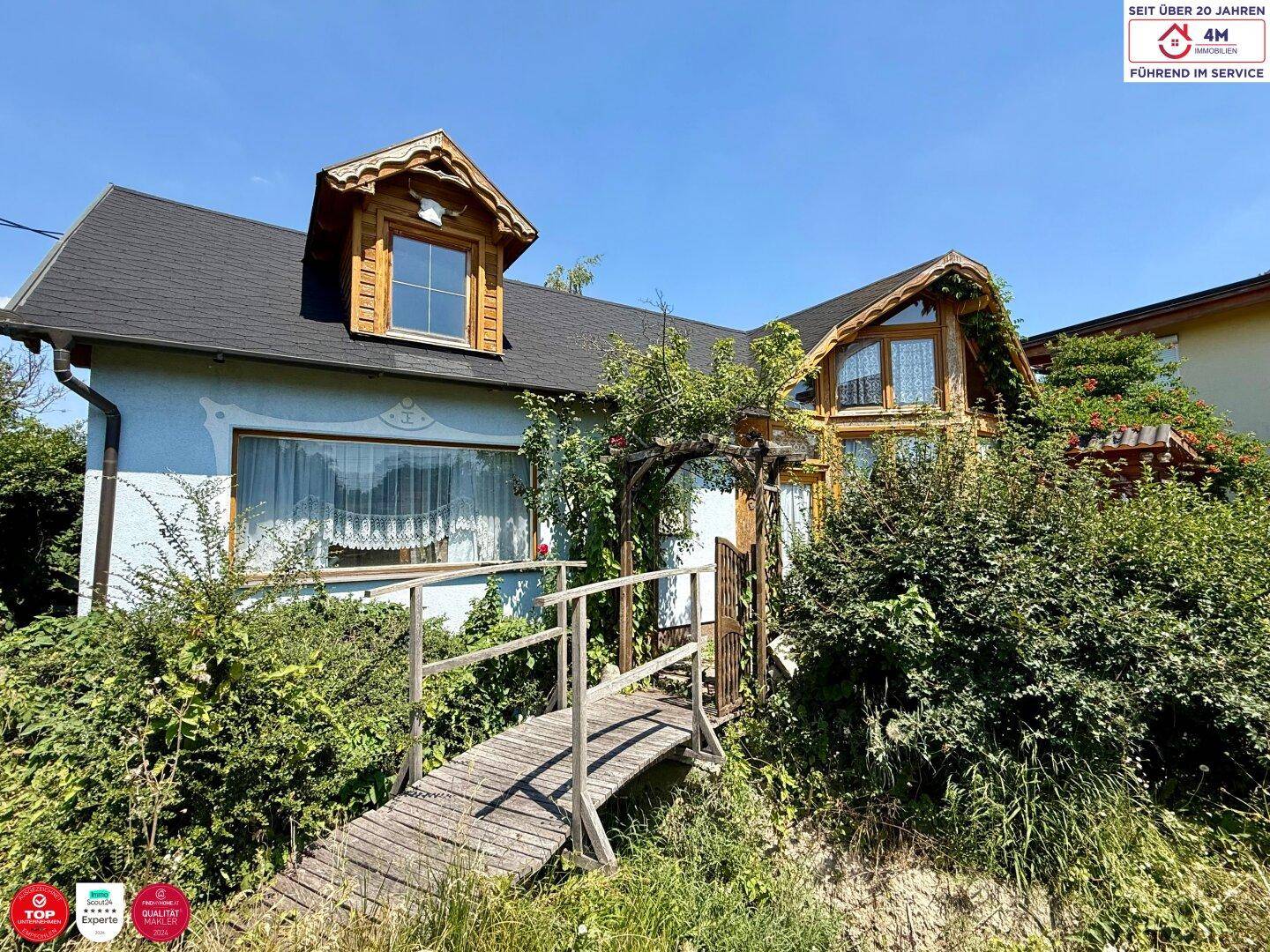 Ansicht des Hauses mit einem kleinen Holzsteg, umgeben von üppigem Grün und blühenden Pflanzen.