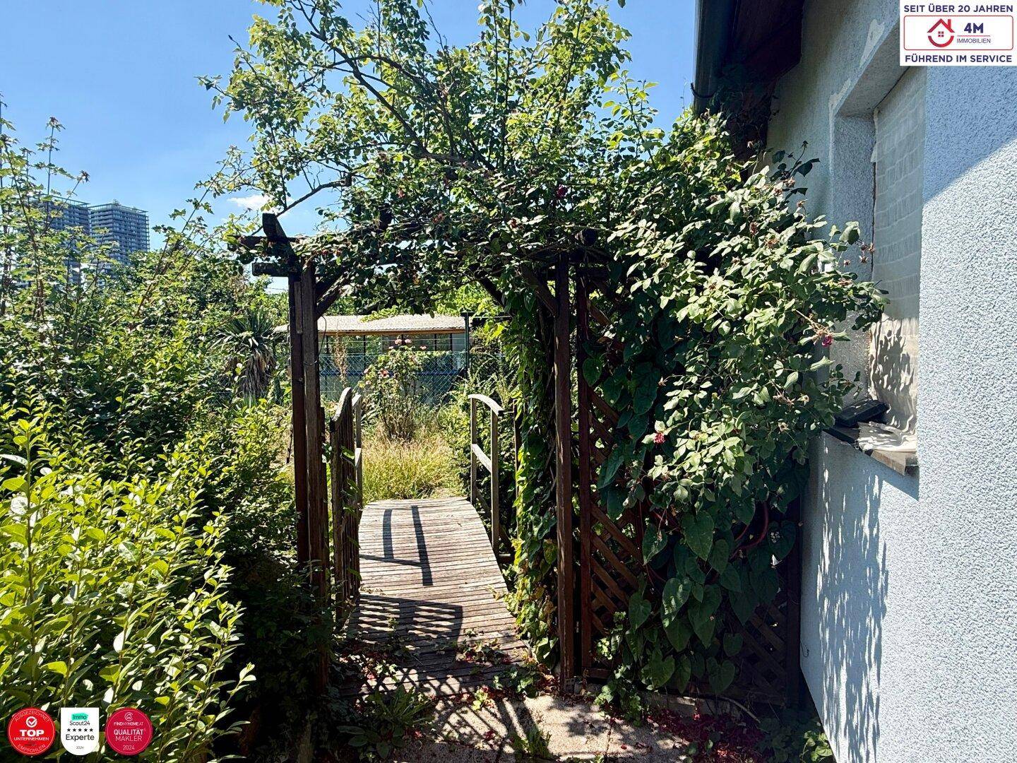 Ein bewachsener Holzbogen führt über einen kleinen Steg in den naturbelassenen Gartenbereich.