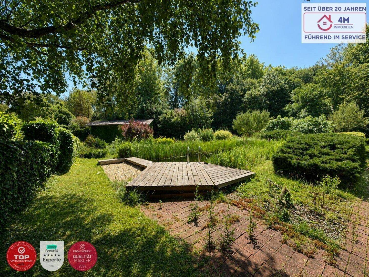 Weitläufiger Gartenbereich mit Teich, Holzsteg und gepflastertem Weg, umgeben von Grün.