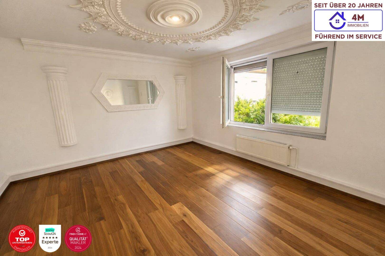 Gemütlicher Raum mit Holzboden, Stuckdecke und großem Fenster mit Rollladen.