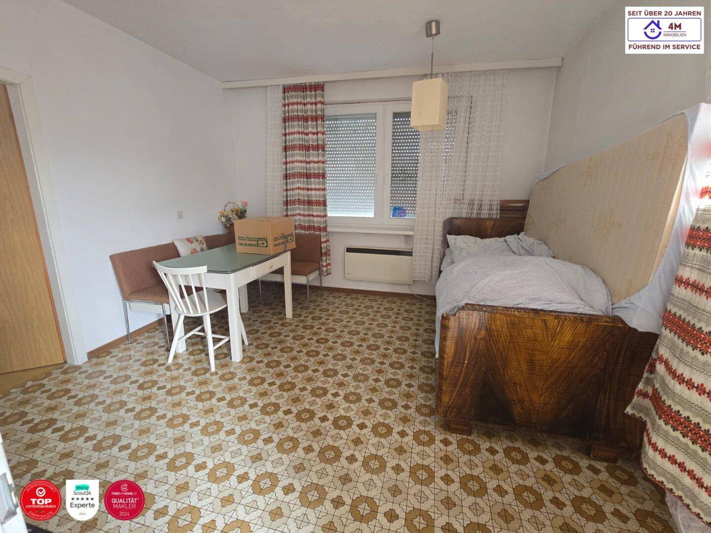 Zimmer mit gemustertem Boden, einem Bett und einem Esstisch mit Stühlen am Fenster.