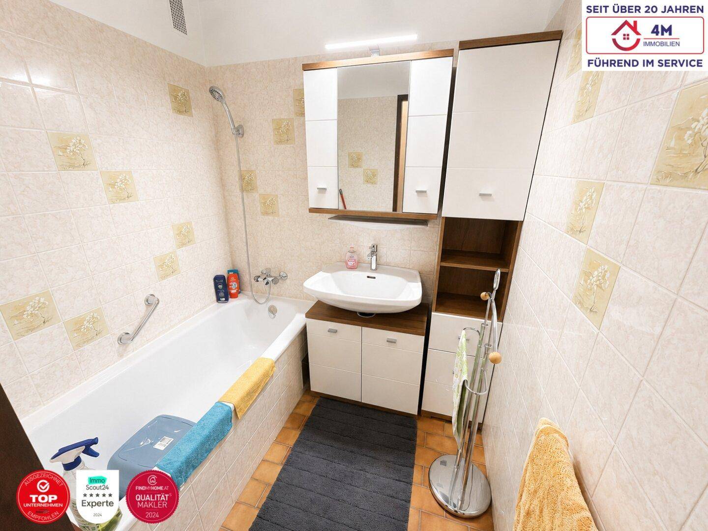 Badezimmer mit Badewanne, Waschbecken und Spiegelschrank, ausgestattet mit hellen Fliesen und Haltegriffen.