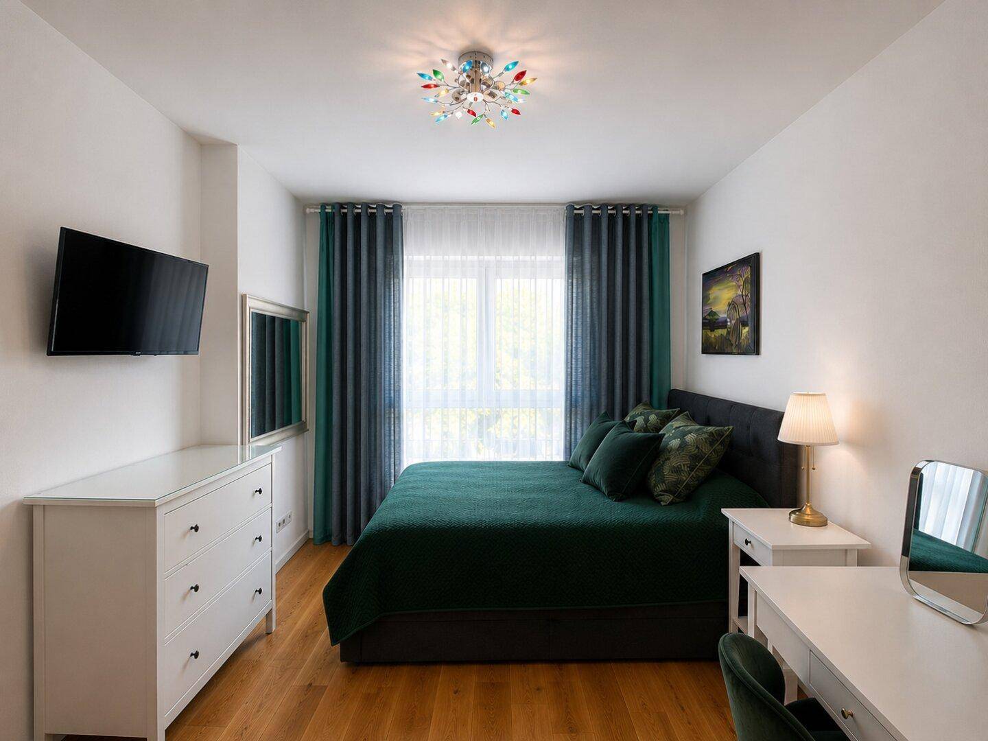 Schlafzimmer mit dunklem Bett, Holzboden, Fernseher und Schreibtisch am Fenster.