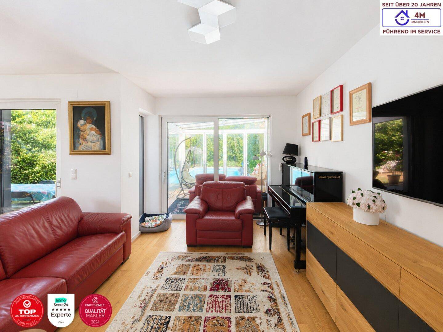 Geräumiges Wohnzimmer mit rotem Sofa, Klavier und Blick in den Wintergarten und Garten.