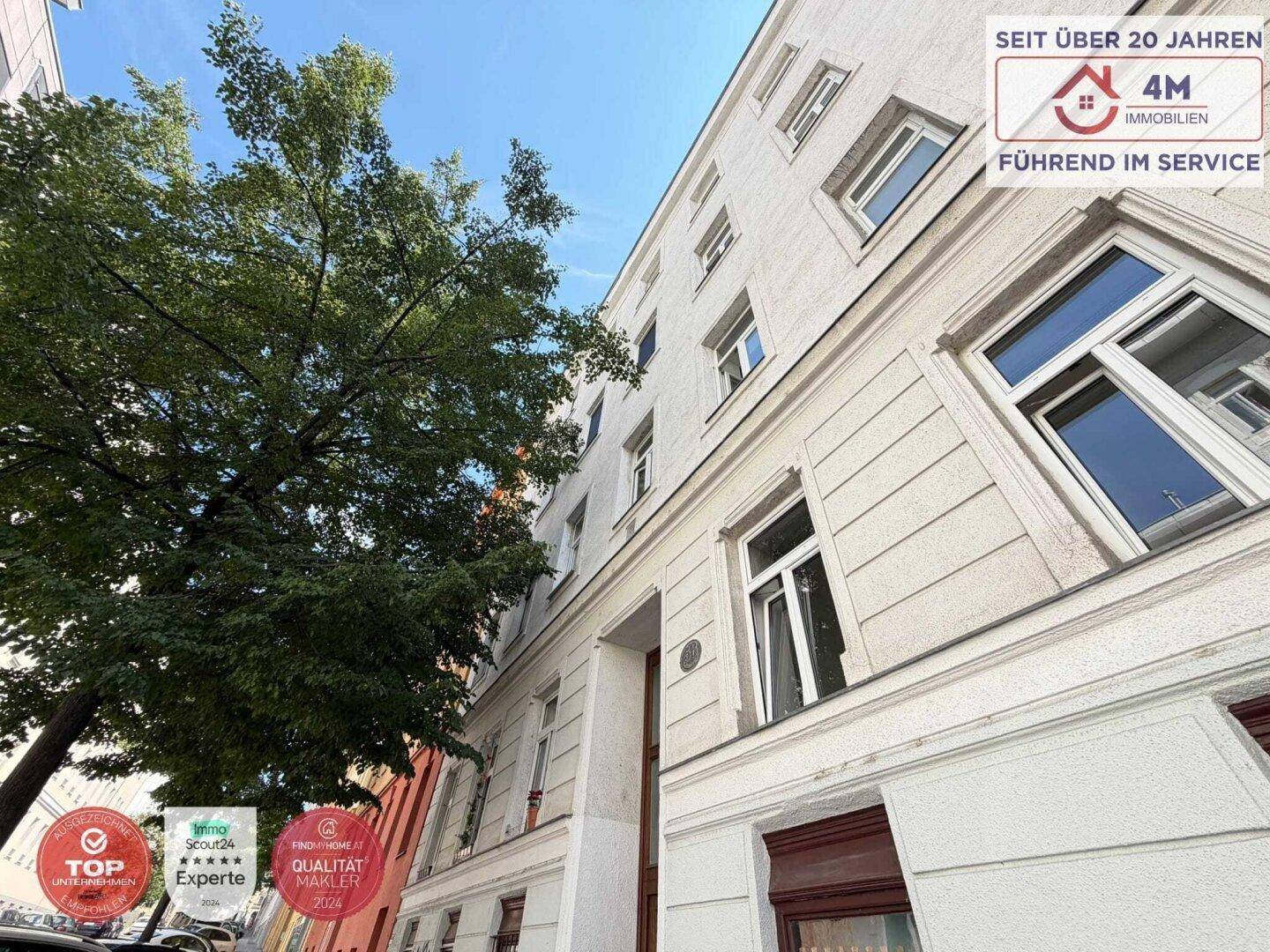 Historische Fassade eines Mehrfamilienhauses mit großen Fenstern und Baumbestand.