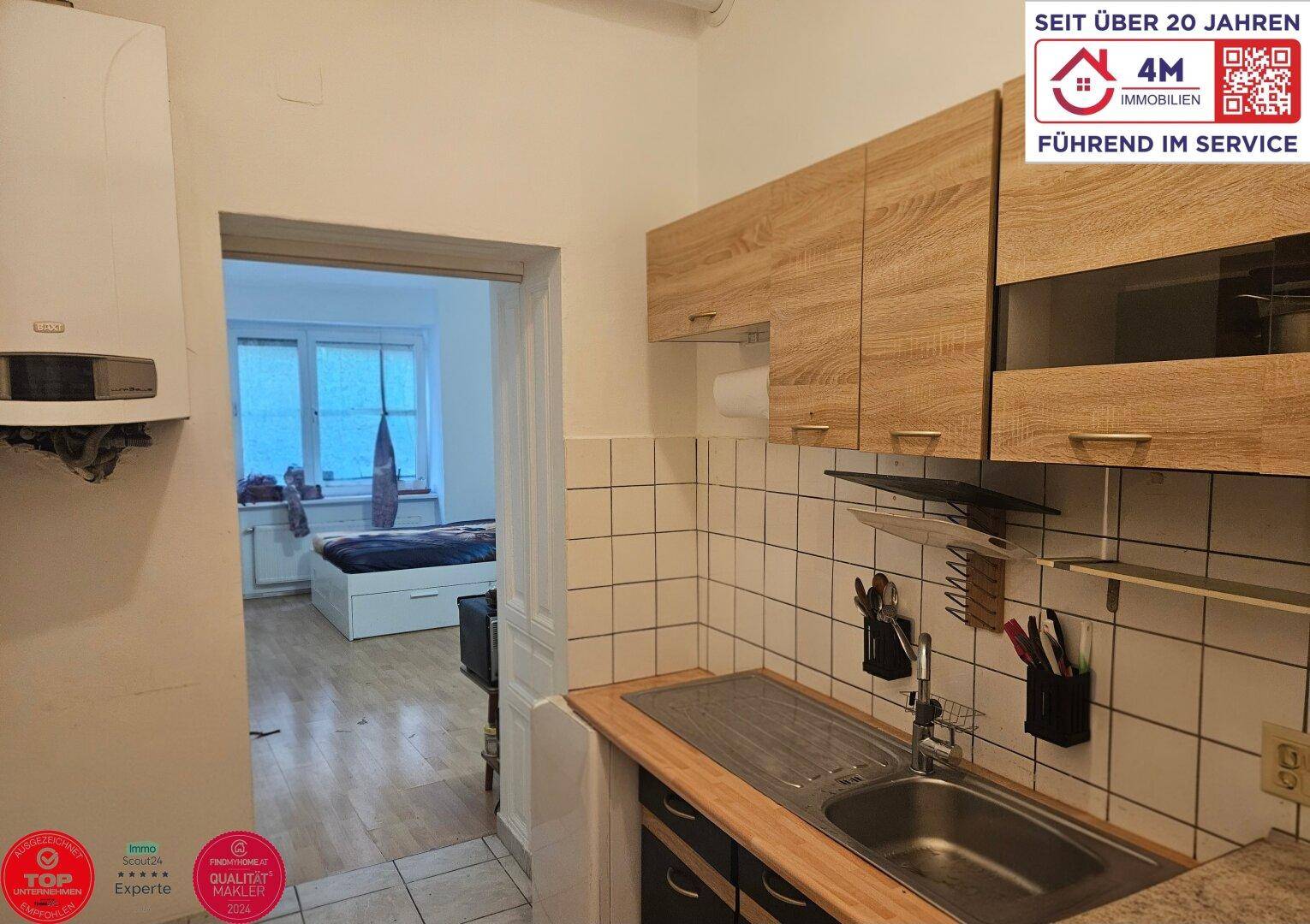 Kleine Küche mit Holzschränken, Spüle und Blick in den angrenzenden Wohn-/Schlafbereich.