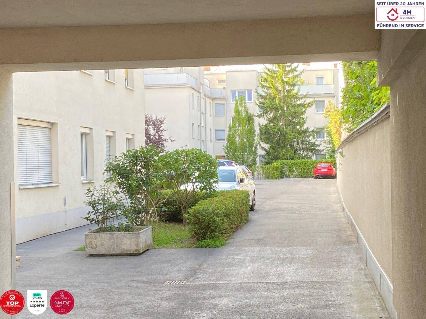 Blick durch einen Torbogen auf einen Innenhof mit Parkplätzen und Bepflanzung.