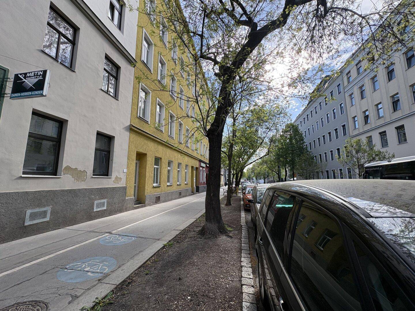 Straßenansicht mit einem gelben Gebäude, Bäumen und parkenden Autos unter blauem Himmel.