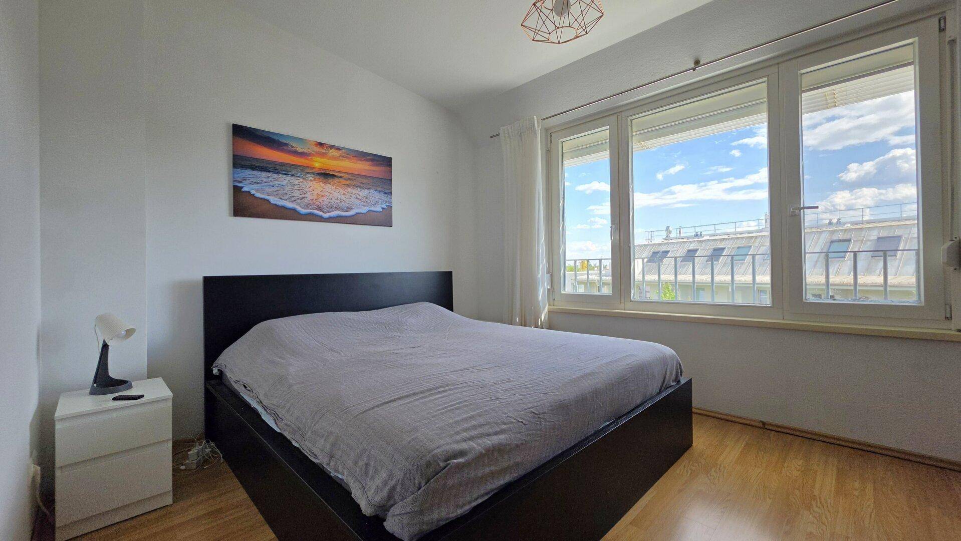 Helles Schlafzimmer mit großem Fenster, Holzboden und Blick auf die Dächer.