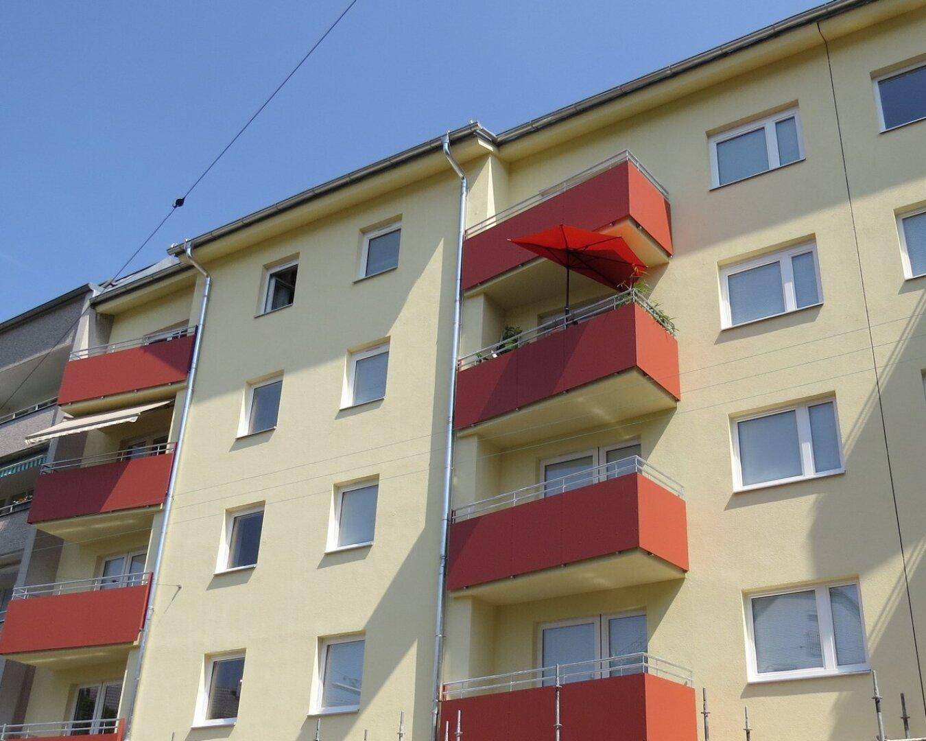Mehrfamilienhaus mit gelber Fassade und roten Balkonen unter blauem Himmel.