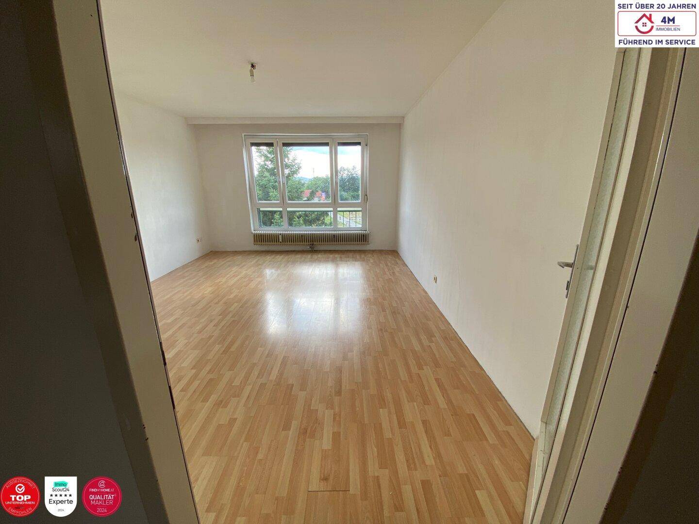 Helles, leeres Zimmer mit Laminatboden und großem Fenster, das viel Tageslicht hereinlässt.