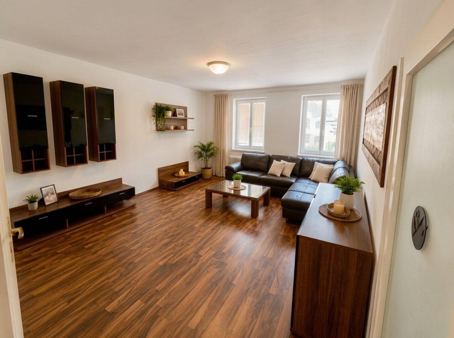 Geräumiges Wohnzimmer mit dunklem Ledersofa, Holzboden und zwei großen Fenstern.