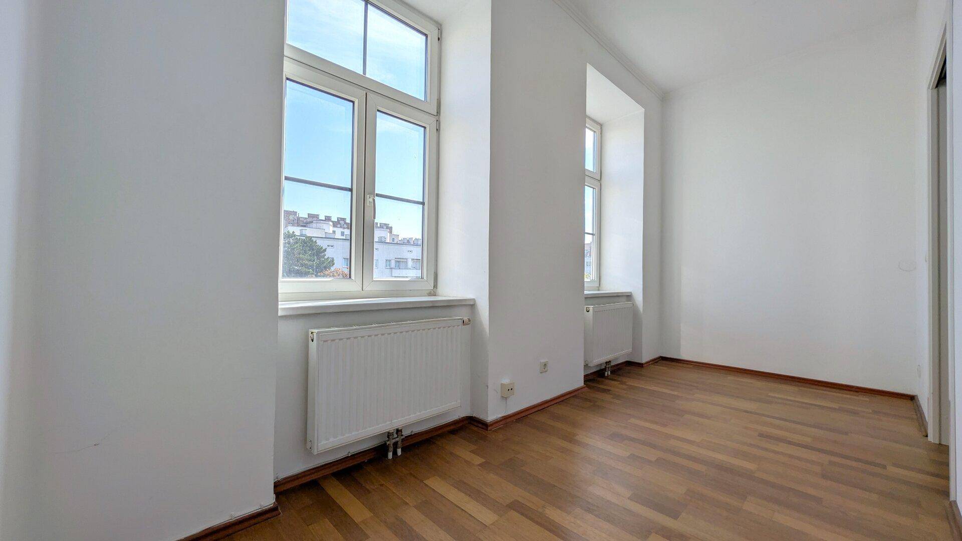 Lichtdurchflutetes Zimmer mit Parkettboden, weißen Wänden und zwei hohen Fenstern.