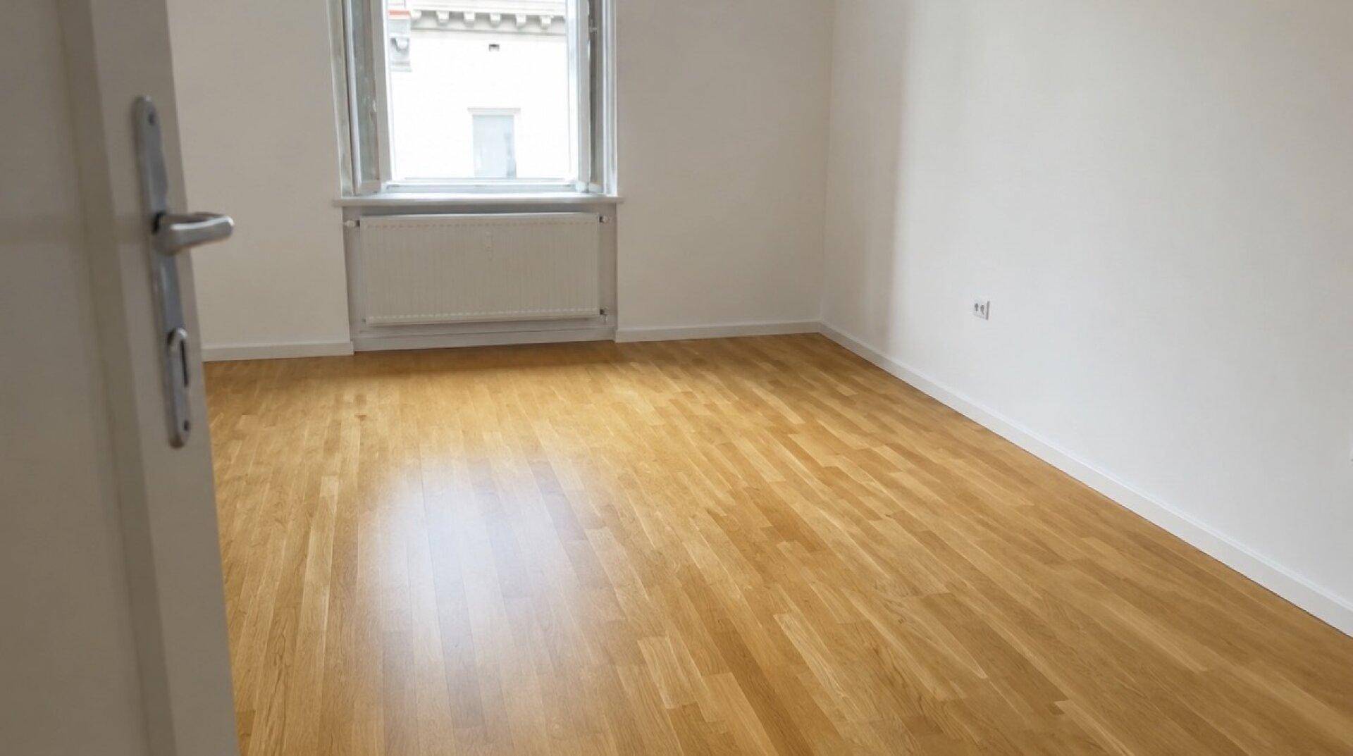 Leeres Zimmer mit hellem Holzboden, weiß gestrichenen Wänden und einem großen Fenster.