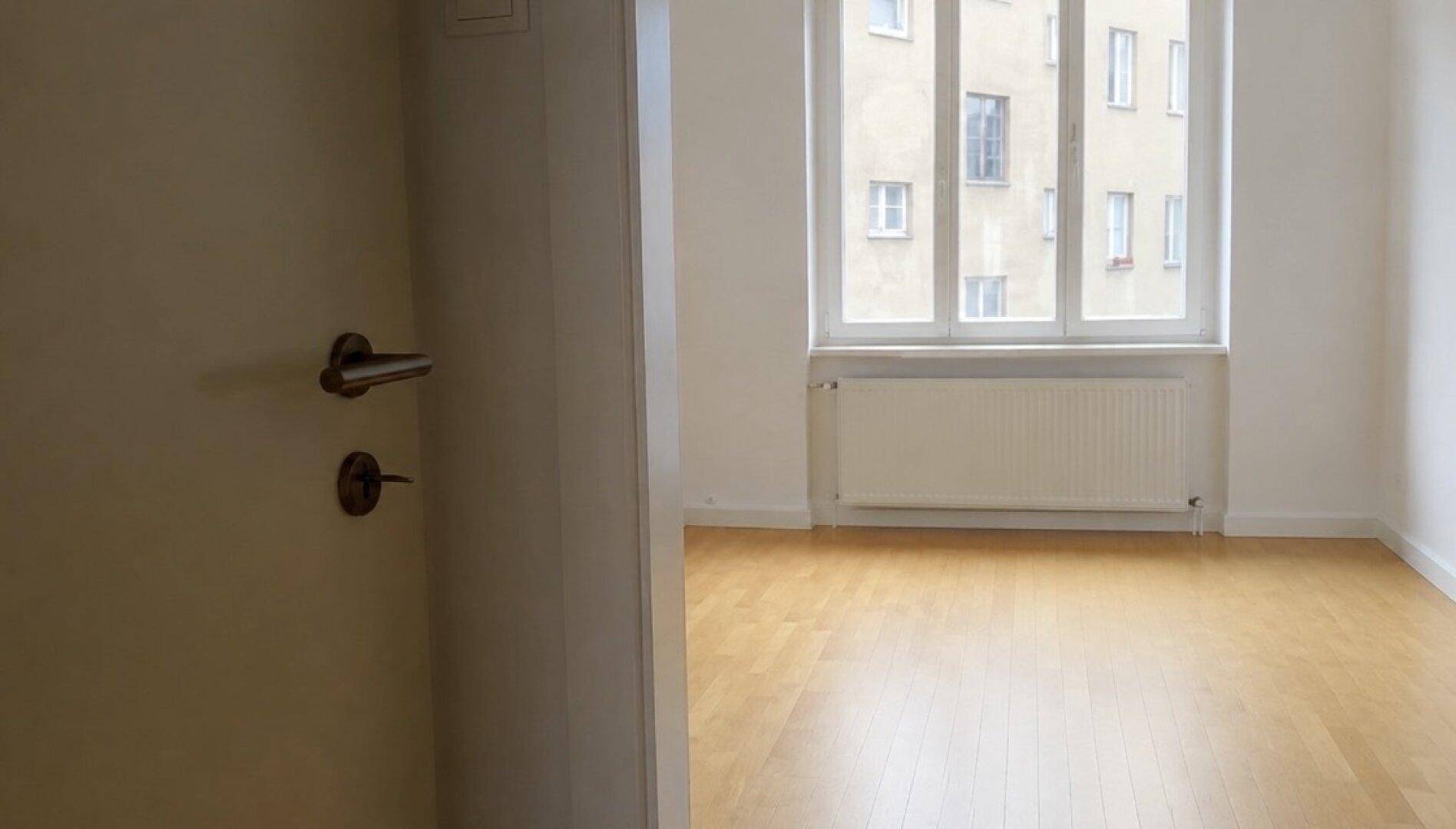 Helles, leeres Zimmer mit Holzboden und Blick auf ein gegenüberliegendes Gebäude.