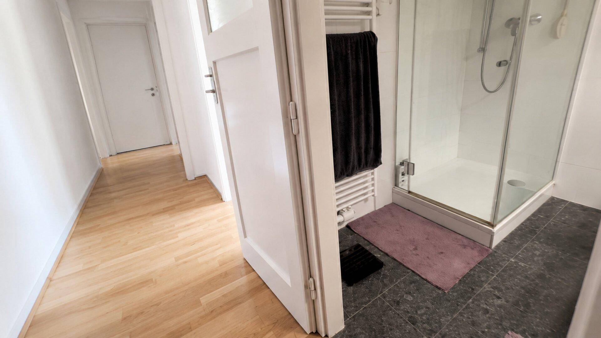 Badezimmer mit Duschkabine und angrenzendem Flur, ausgestattet mit einem Handtuchheizkörper.