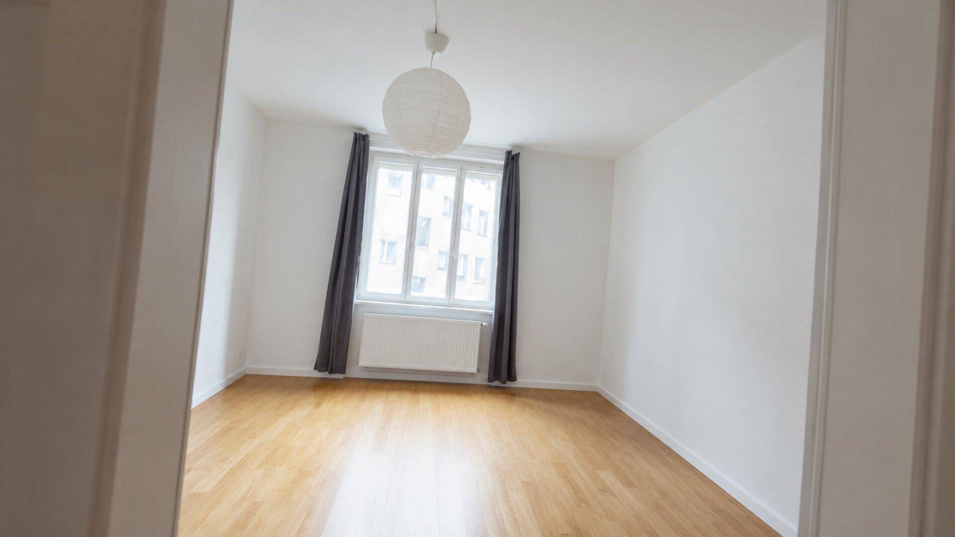 Leeres Zimmer mit Holzboden, weißen Wänden und einem Fenster mit dunklen Vorhängen.