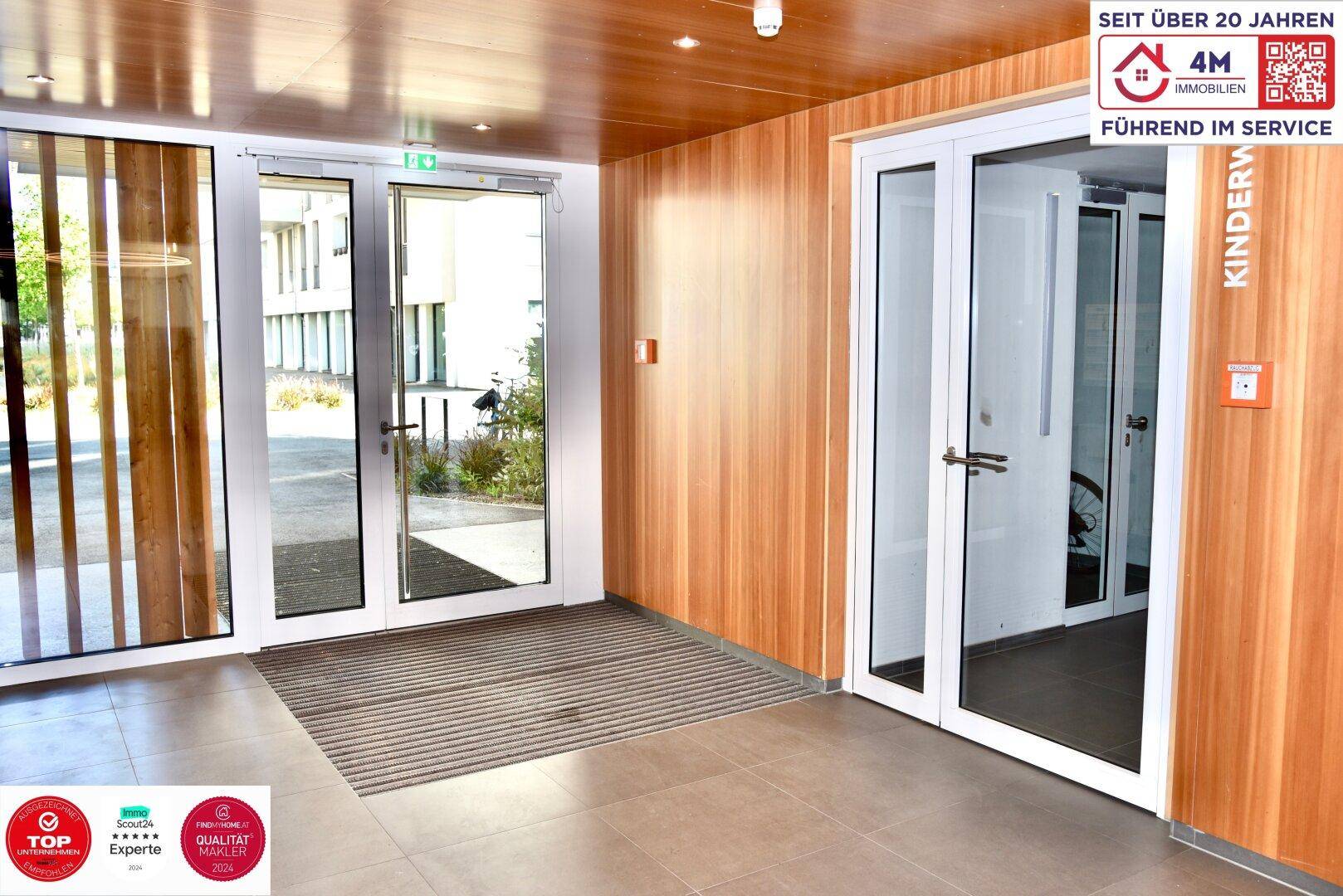 Heller Eingangsbereich des Gebäudes mit großen Glastüren und Holzverkleidung, modern und einladend.