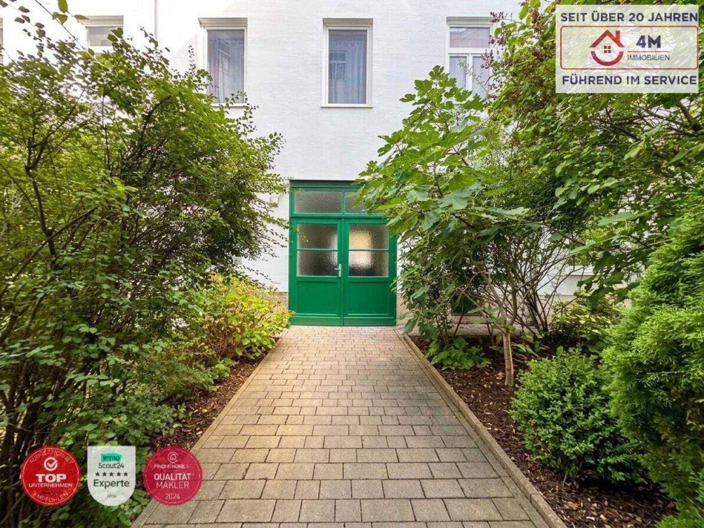 Gepflasterter Weg führt zum grünen Eingang einer Wohnanlage, umgeben von gepflegten Sträuchern.