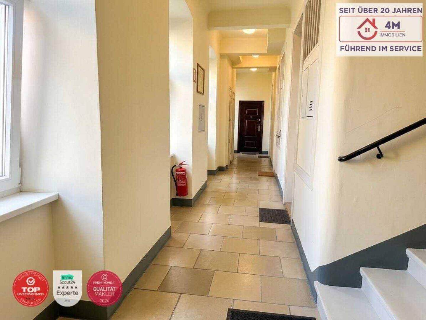 Langer, heller Flur mit Fliesenboden, Treppenaufgang und einer dunklen Eingangstür.