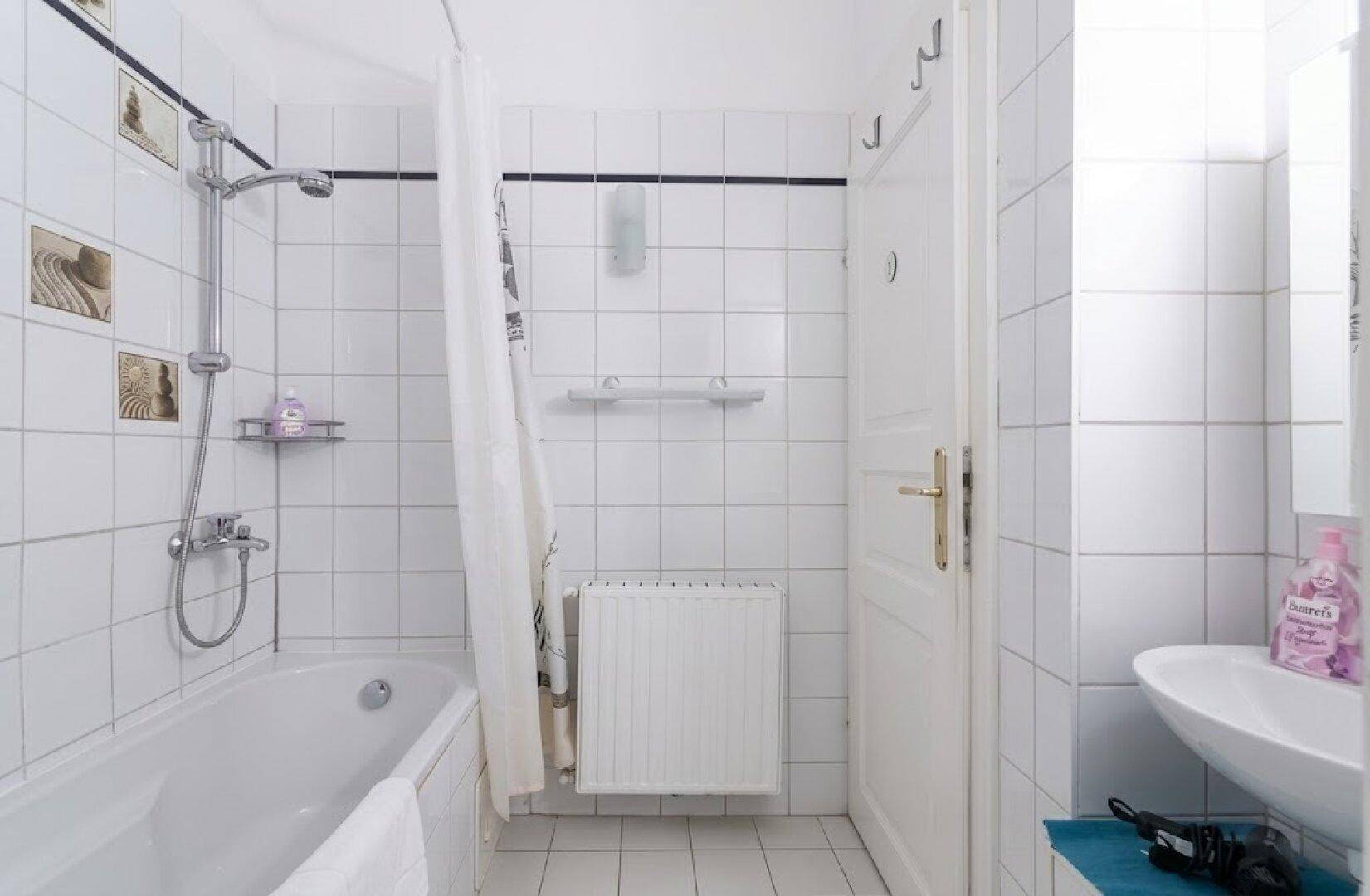 Helles Badezimmer mit weißen Fliesen, Badewanne mit Duschfunktion und einem Waschbecken.