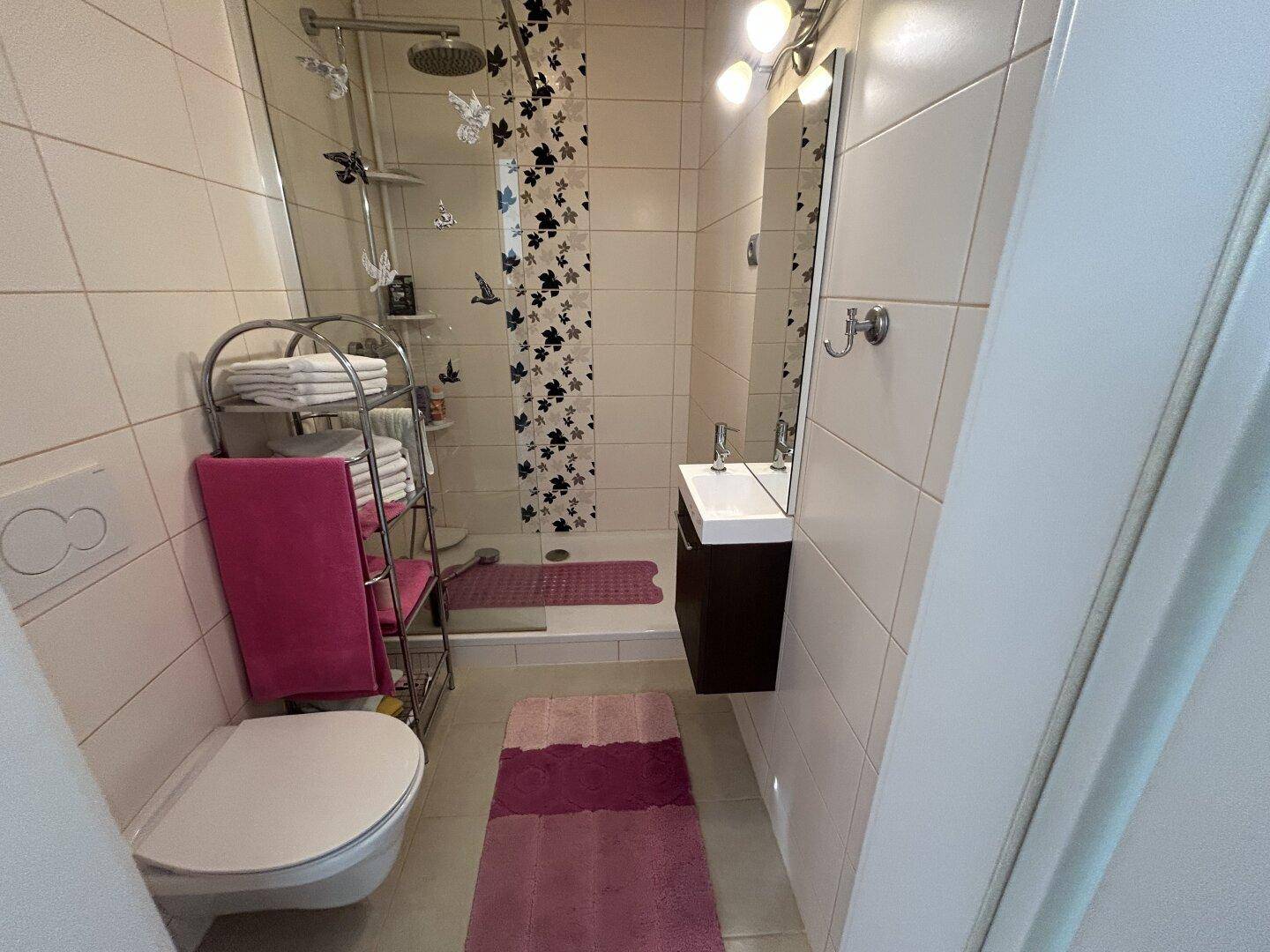 Modernes Badezimmer mit Duschkabine, Hänge-WC und kleinem Waschbecken in hellen Fliesen.
