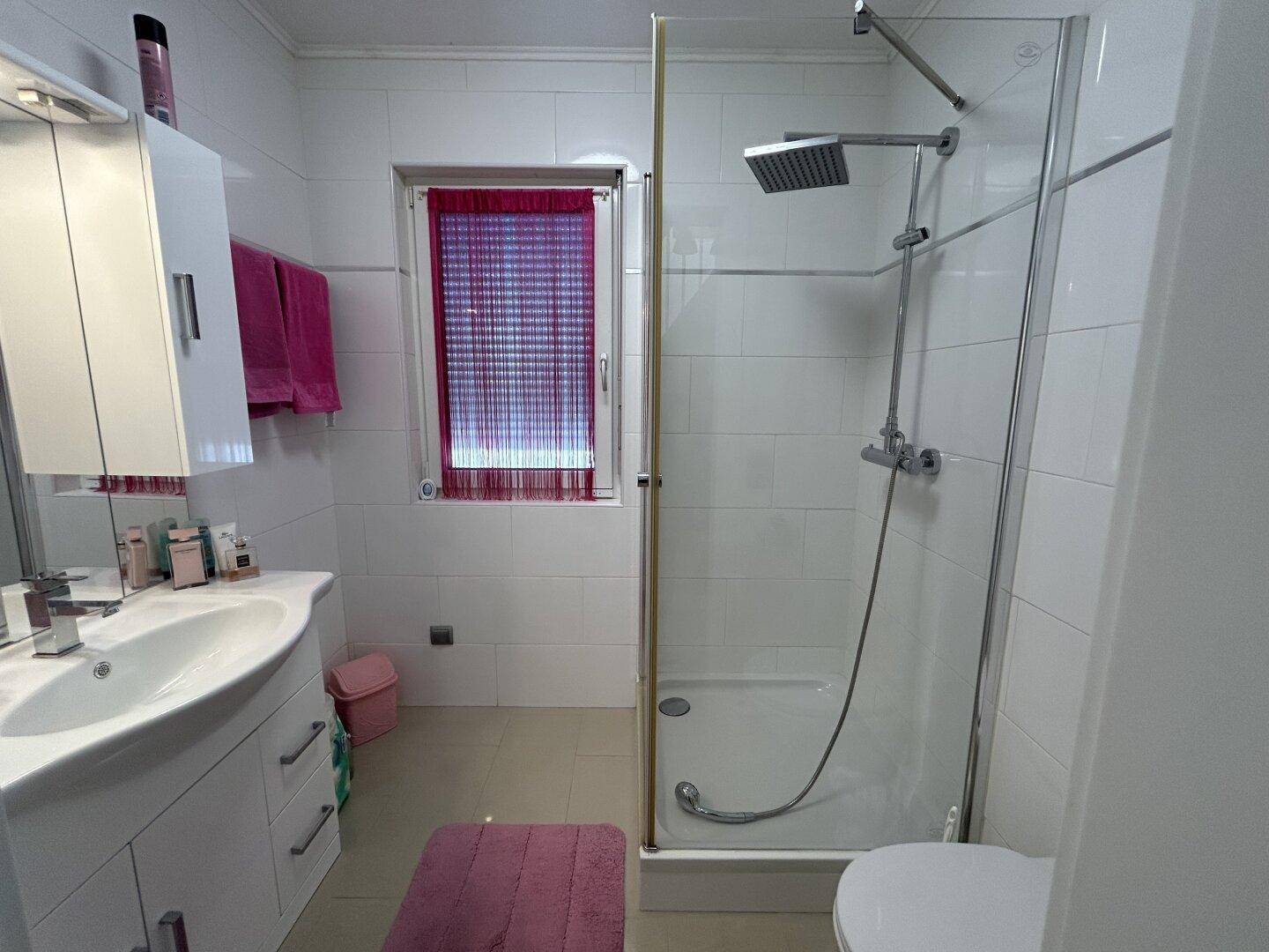 Helles Badezimmer mit Duschkabine, Waschbecken und Fenster, ausgestattet mit weißen Fliesen.