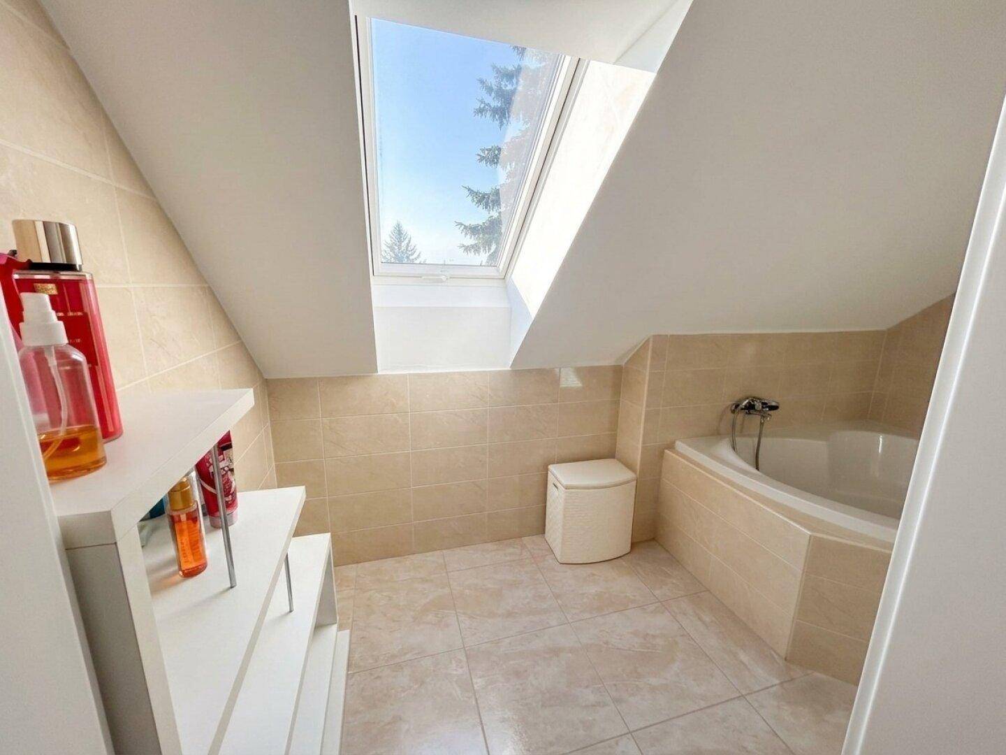 Helles Badezimmer im Dachgeschoss mit Badewanne, beigen Fliesen und großem Dachfenster für Tageslicht.