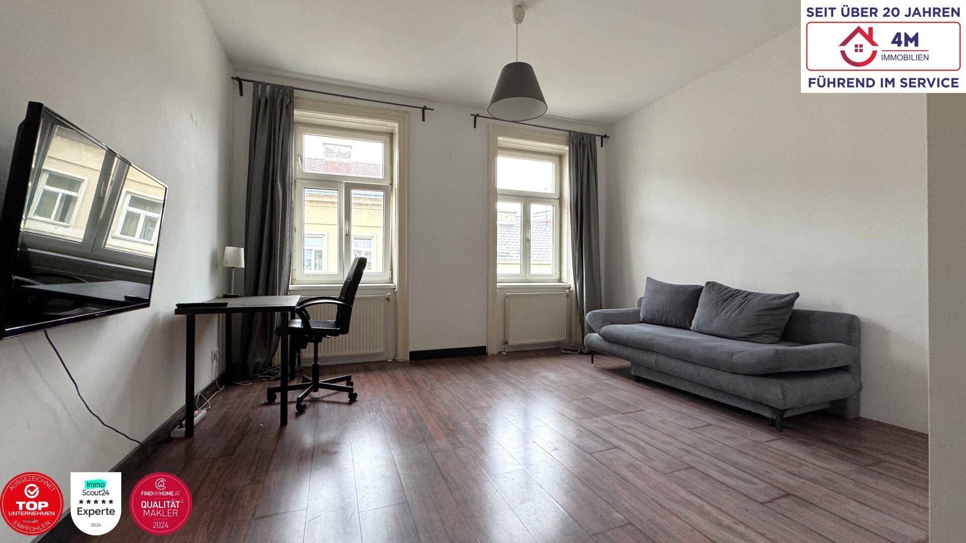 Geräumiges Wohnzimmer mit Sofa, Schreibtisch und zwei großen Fenstern, die viel Licht hereinlassen.