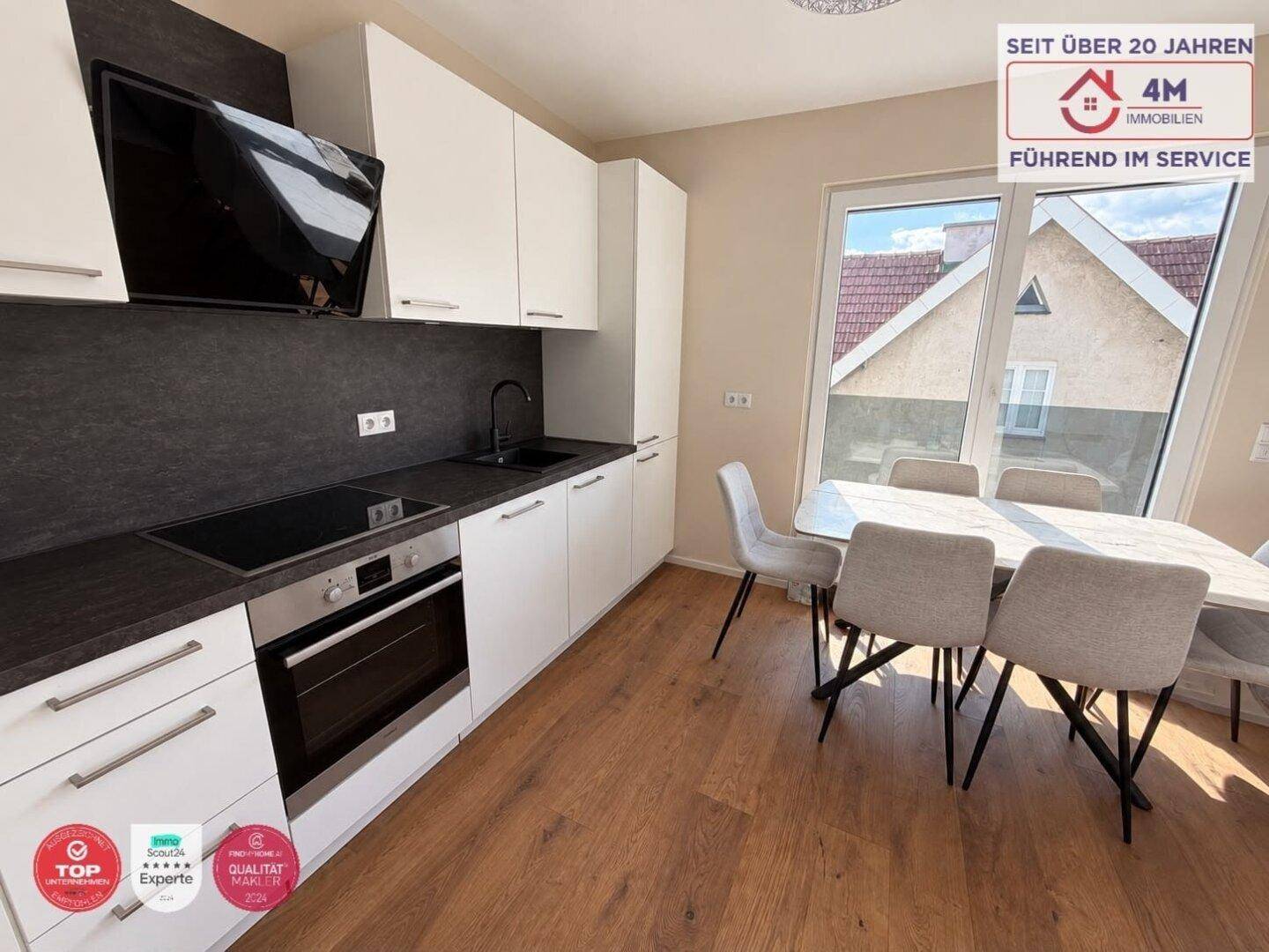 Moderne Einbauküche mit weißen Schränken, dunkler Arbeitsplatte und Essbereich am Fenster.