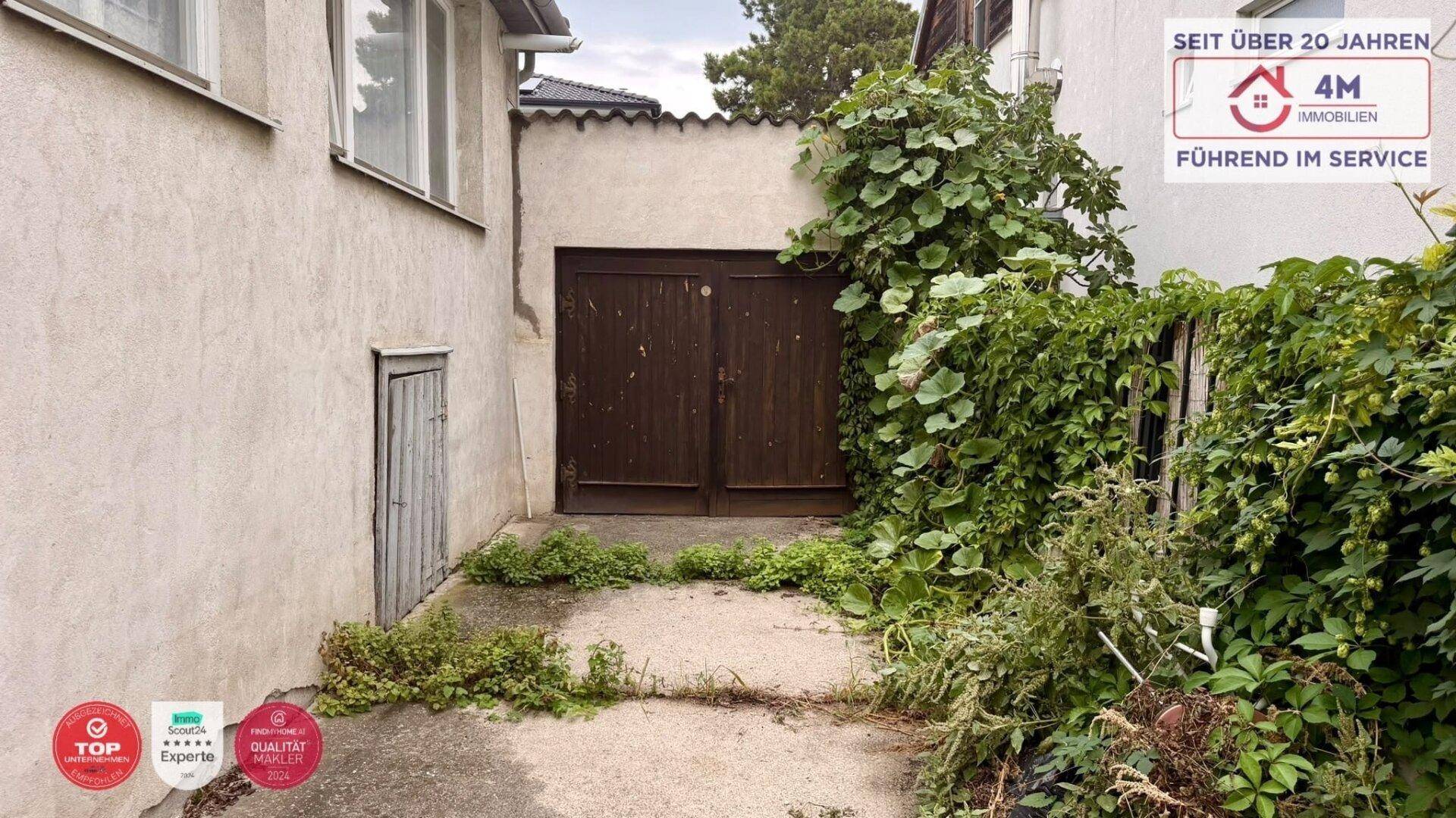 Alte Garage mit Holztüren und stark bewachsenem Eingangsbereich des Grundstücks.
