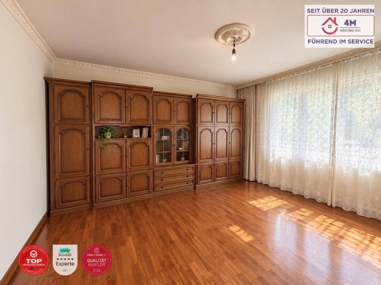 Wohnzimmer mit großem Holzschrank, Parkettboden und viel Tageslicht durch das Fenster.