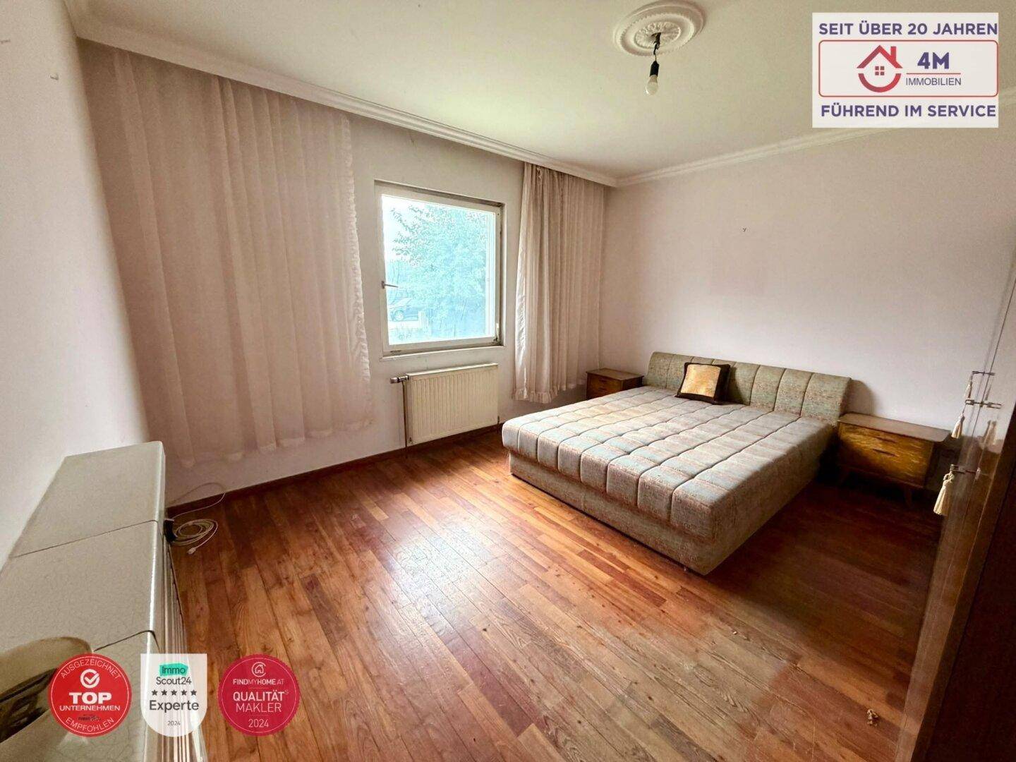 Schlafzimmer mit Parkettboden, Bett und Fenster mit Blick ins Grüne.