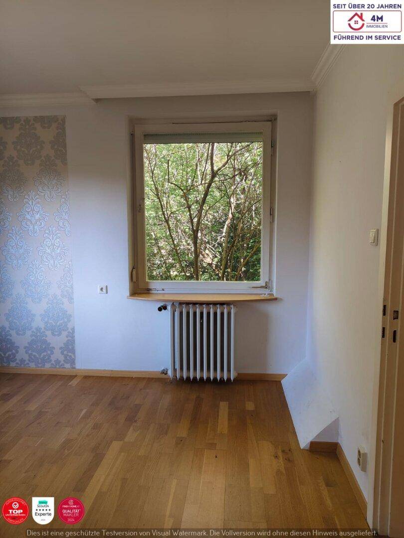 Ein heller Raum mit Parkettboden, Heizkörper und Fenster mit Blick ins Grüne.
