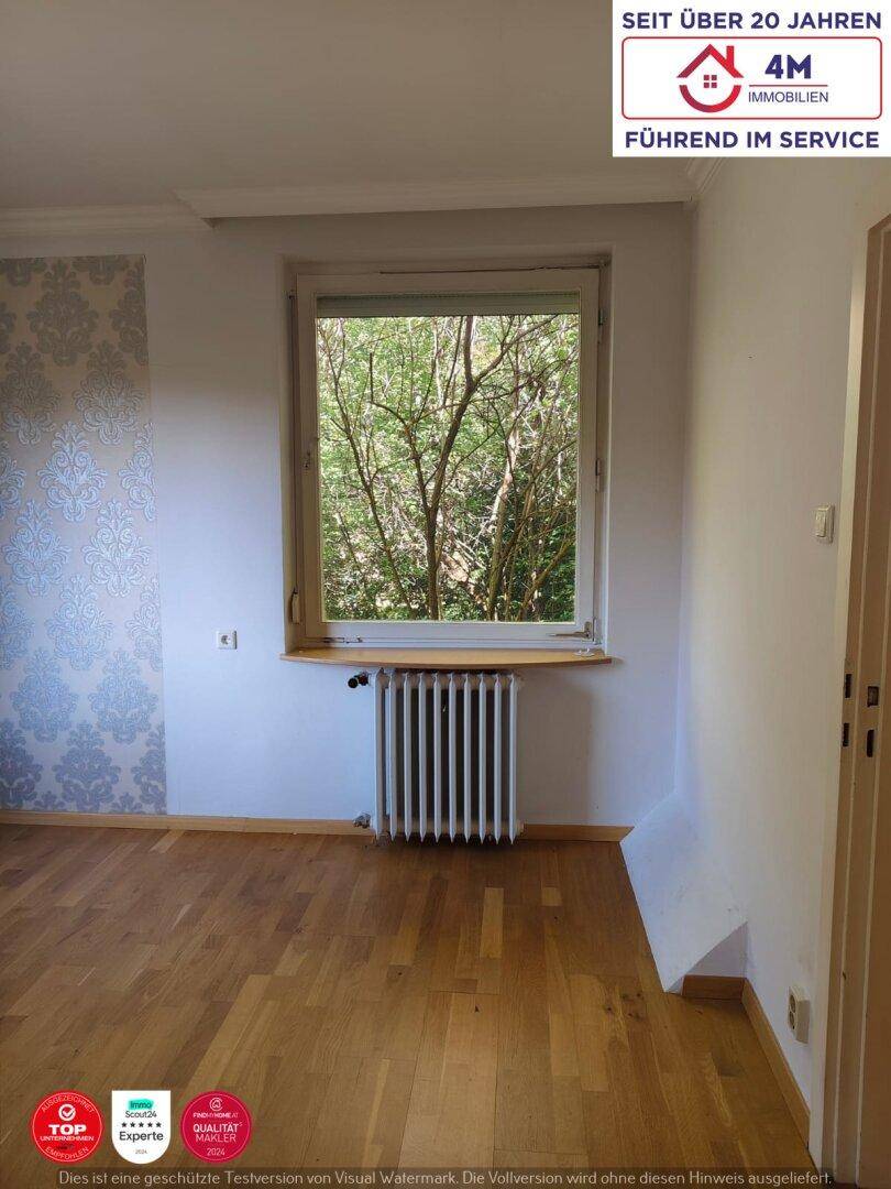 Ein heller Raum mit Parkettboden, Heizkörper und Fenster mit Blick ins Grüne.